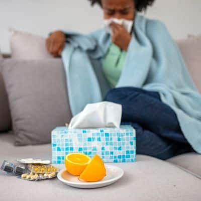 woman on couch holding tissue to nose medicine and orange on coffee table